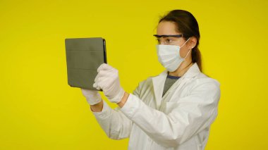 Tıbbi maskeli, gözlüklü ve lateks eldivenli genç doktor tablete bakıyor.