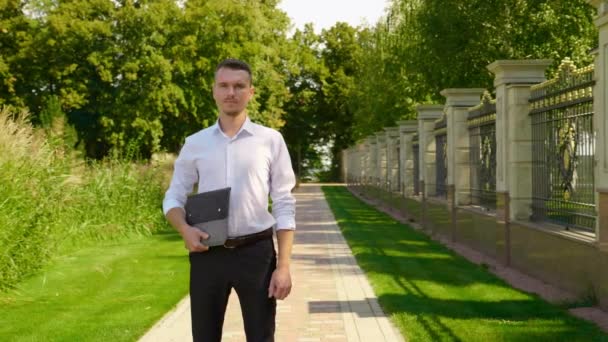 Geschäftsmann steht mit Laptop in der Hand in einem Park und blickt in die Kamera — Stockvideo