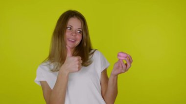 Beyaz tişörtlü bir kadın pembe bir badem kurabiyesiyle elini kaldırıyor ve gülümsüyor.