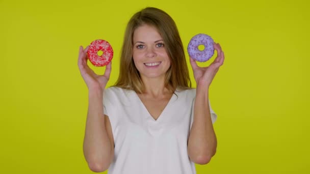 Mulher levanta as mãos com donuts azuis e vermelhos, sorrindo em um fundo amarelo — Vídeo de Stock