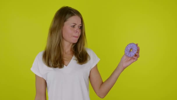 Woman looks at the blue donuts in hand, licks, bites lips on a yellow background — Stock Video