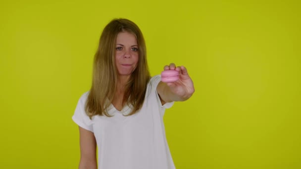 Woman in a white Tshirt looks at the pink macaroon in hand, licks and bites lips — Stock Video