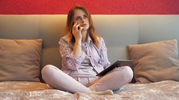 Жінка сидить на ліжку з планшетом в руці і розмовляє по телефону — стокове відео
