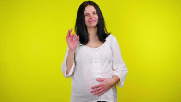 Έγκυος γυναίκα κρατά το χέρι στο στομάχι, δείχνει εντάξει και χαμογελά σε ένα κίτρινο φόντο — Αρχείο Βίντεο