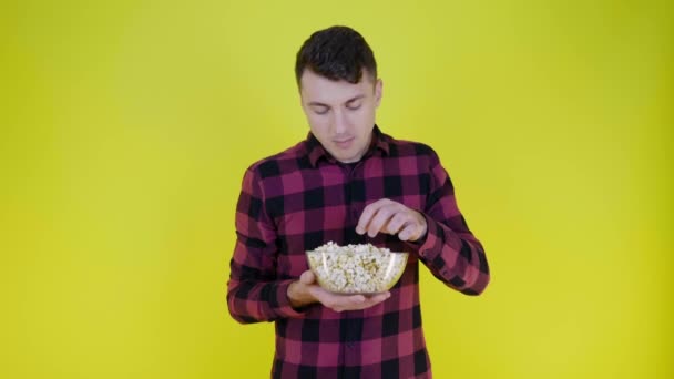 Man in een roze geruite shirt eet popcorn uit glazen schaal op gele achtergrond — Stockvideo