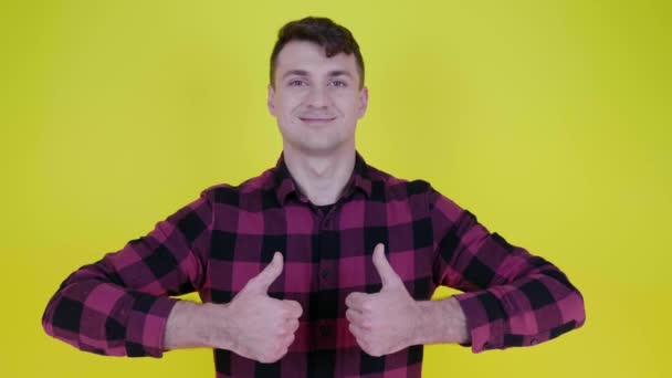 Man in roze geruite shirt glimlacht, heft handen en toont als op gele achtergrond — Stockvideo