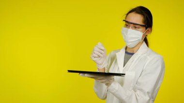 Tıbbi maskeli, gözlüklü ve lateks eldivenli doktor elinde bir tablet tutuyor.