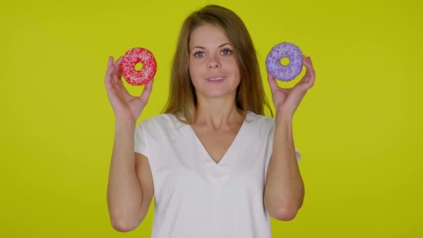 Mulher levanta as mãos com donuts azuis e vermelhos, sorrindo em um fundo amarelo — Vídeo de Stock