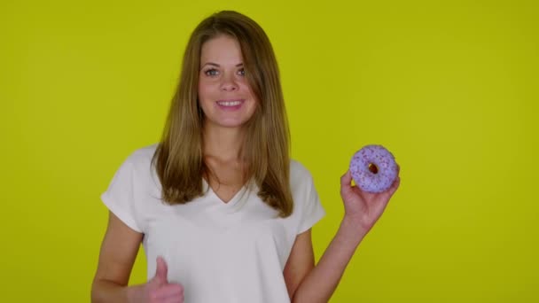 Mulher de t-shirt branca levanta a mão com um donuts azul, mostra como, sorri — Vídeo de Stock