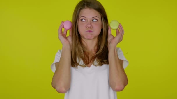 Γυναίκα κοιτάζει ένα ροζ, κίτρινο αμυγδαλωτά στα χέρια με pouting μάγουλα, το στόμα — Αρχείο Βίντεο