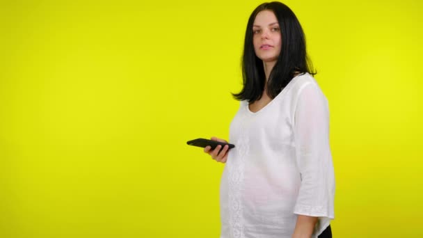 Jeune femme enceinte tient un smartphone en main et regarde la caméra — Video