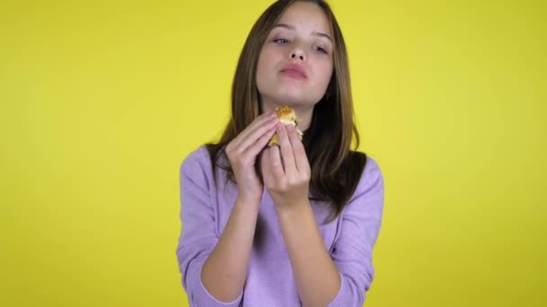 Tonåring flicka i en rosa tröja biter och äter hamburgare på gul bakgrund — Stockvideo