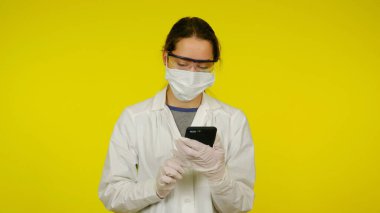 Tıbbi maskeli, gözlüklü ve lateks eldivenli doktor akıllı telefona bakar.