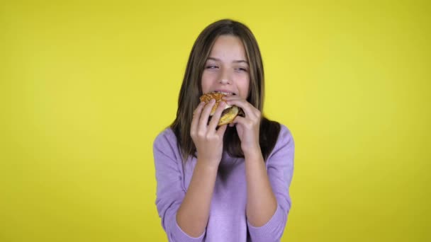 Teen κορίτσι σε ένα ροζ πουλόβερ τρώει ένα burger σε κίτρινο φόντο με αντίγραφο χώρο — Αρχείο Βίντεο