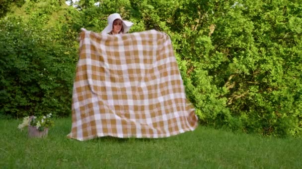 Donna diffonde un plaid su erba in un giardino botanico sullo sfondo di alberi — Video Stock