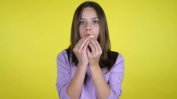 Menina adolescente em uma camisola rosa morde e come hambúrguer no fundo amarelo — Vídeo de Stock