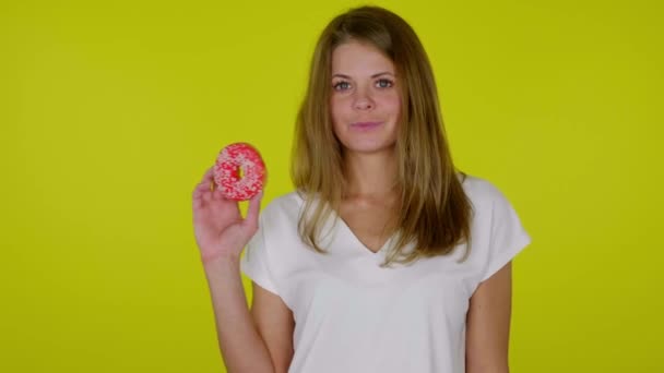 Mulher levanta a mão com um donuts vermelho, mostra como, sorri em um fundo amarelo — Vídeo de Stock