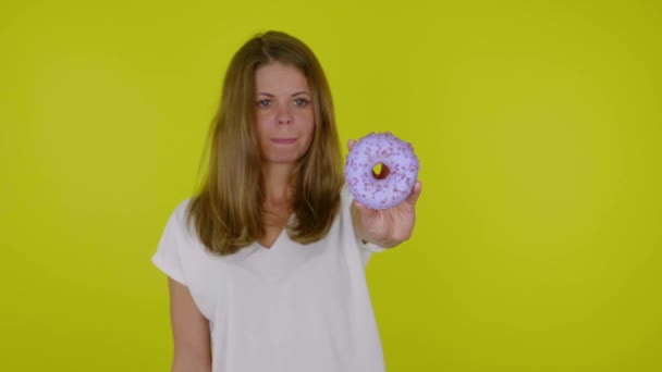 Mulher em uma camiseta branca olha para os donuts azuis na mão, lambe e morde os lábios — Vídeo de Stock
