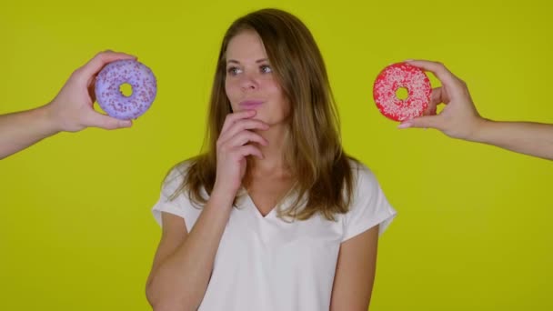 Mulher positiva em uma camiseta olha em volta, sorri e escolhe qual donut comer — Vídeo de Stock