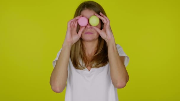 Rolig kvinna i vit T-shirt håller gula, rosa makaroner i händerna, grimaser — Stockvideo