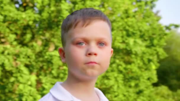 Liten pojke i en vit t-shirt ser sig omkring i parken på en bakgrund av träd — Stockvideo