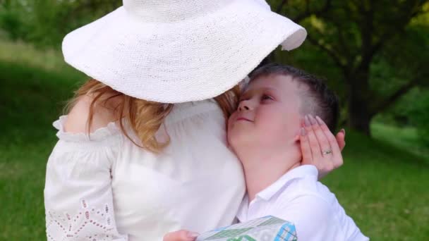 Anne şapkalı ve oğlu parkta çimenlerde oturuyor, sarılıyor, öpüşüyor, gülümsüyor. — Stok video