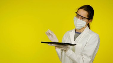 Tıbbi maskeli, gözlüklü ve lateks eldivenli doktor elinde bir tablet tutuyor.