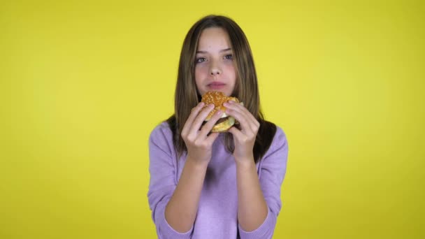 Девушка в розовом свитере кусает и ест бургеры на желтом фоне. — стоковое видео