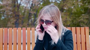 Güneş gözlüklü bir kadın cep telefonuyla konuşuyor, açık hava parkında bankta oturuyor.