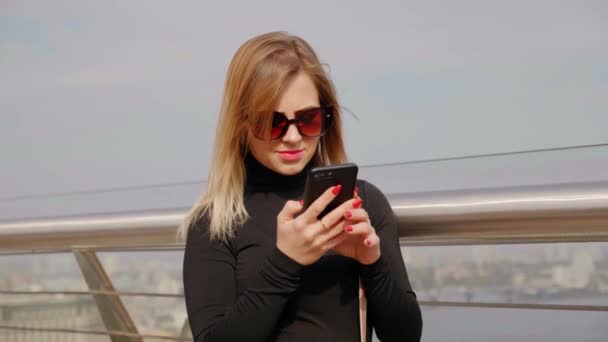 Mujer en gafas de sol y cuello alto negro utiliza teléfono inteligente al aire libre — Vídeos de Stock