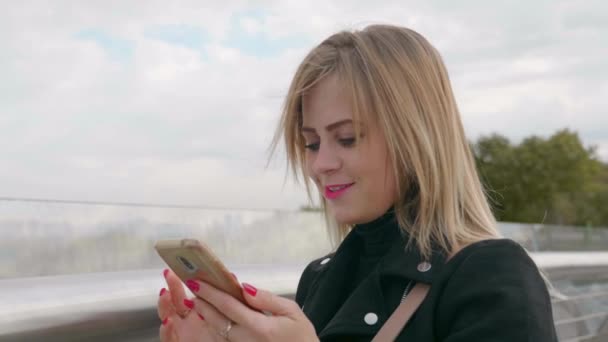 Mulher positiva em uma jaqueta preta usa smartphone ao ar livre e sorrisos — Vídeo de Stock
