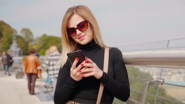 Mulher positiva em óculos de sol, sorrisos de gola alta preta, usa smartphone ao ar livre — Vídeo de Stock