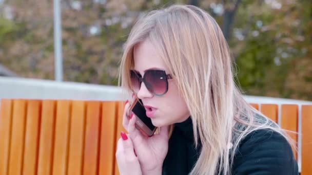 Gros plan femme dans les lunettes de soleil parler sur le téléphone portable, assis sur le banc extérieur — Video