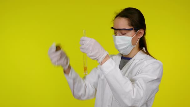 Mujer con abrigo médico, máscara protectora y gafas gotea reactivo en el tubo de ensayo — Vídeos de Stock