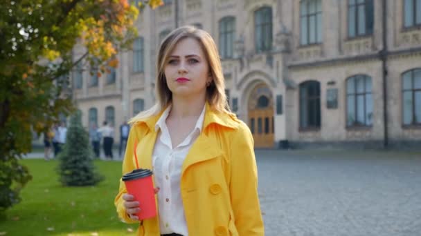 Nő fehér blúzban, sárga köpeny kávéval a kezében sétál a parkban. — Stock videók