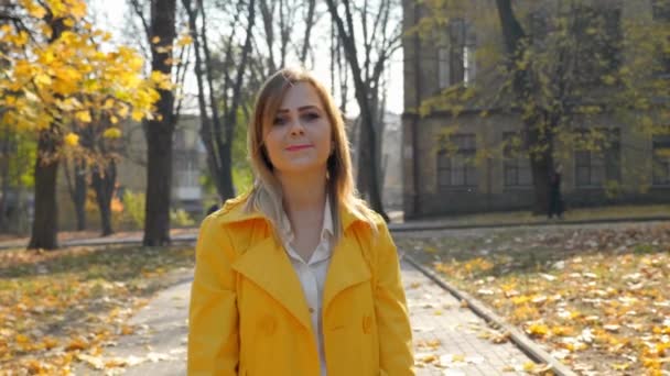 Vreugdevolle vrouw gele mantel loopt herfst park, kijkt naar de camera en glimlacht — Stockvideo