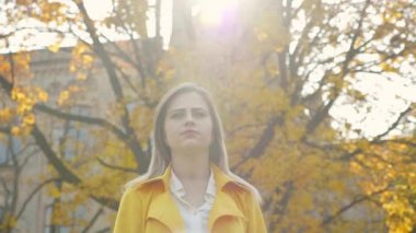 Pelerinli bir kadın, beyaz bluzlu, arka plandaki ağaçta yürüyen sarı yapraklı bir kadın.