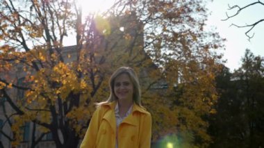 Pelerinli kadın, beyaz bluz arka planda duruyor sarı yapraklı, gülümseyen.