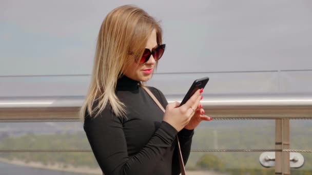 Женщина в солнечных очках и черной водолазке пользуется смартфоном на открытом воздухе — стоковое видео