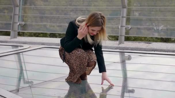 Kvinna i svart jacka, leopardkjol sitter på glasbro, tittar ner utomhus — Stockvideo
