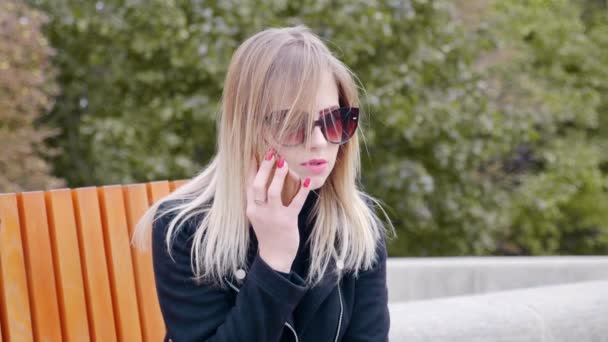 Femme en lunettes de soleil parlant sur un téléphone portable, assise sur un banc dans un parc extérieur — Video