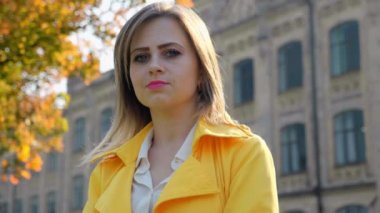 Beyaz bluzlu ve sarı pelerinli bir kadın dışarıdaki kameraya bakıyor. Yavaş çekim