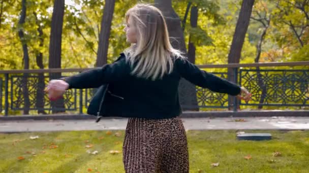 Lycklig kvinna svart jacka, leopard kjol promenader i parken, snurrar med glädje — Stockvideo