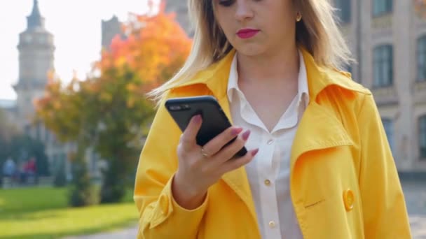 Närbild kvinna i vit blus och gul mantel använder smartphone och promenader park — Stockvideo