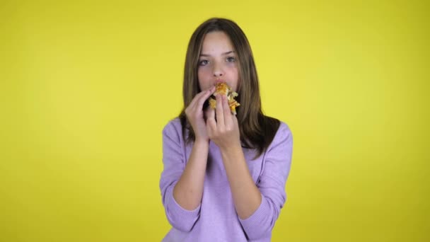 Adolescente chica en un suéter rosa muerde y come hamburguesa sobre fondo amarillo — Vídeo de stock