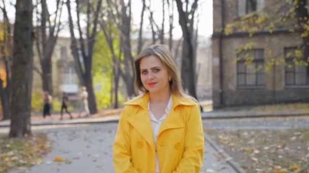 Žena ve žlutém plášti, bílá blůza, úsměvy, stojí podzimní park, vypadá kamerou — Stock video