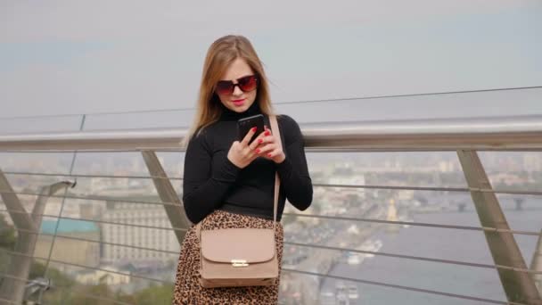 Donna in occhiali da sole, dolcevita nero, borsa marrone utilizza smartphone all'aperto — Video Stock