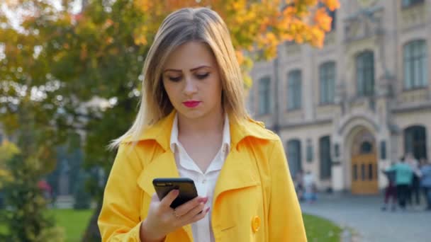 Femme en chemisier blanc, manteau jaune utilise smartphone, promenades parc dactylographie message — Video