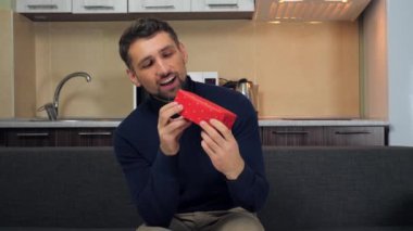 Mavi kazaklı mutlu adam kırmızı kutuyu inceliyor, sarılıyor, hediyesini veriyor, gülümsüyor.