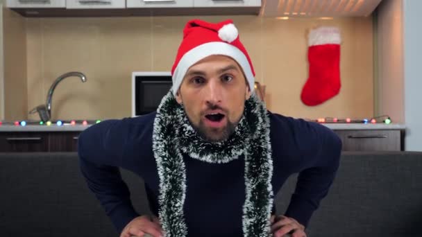 Geschokt man in Nieuwjaar rood Kerstman hoed kijkt televisie, open mond — Stockvideo